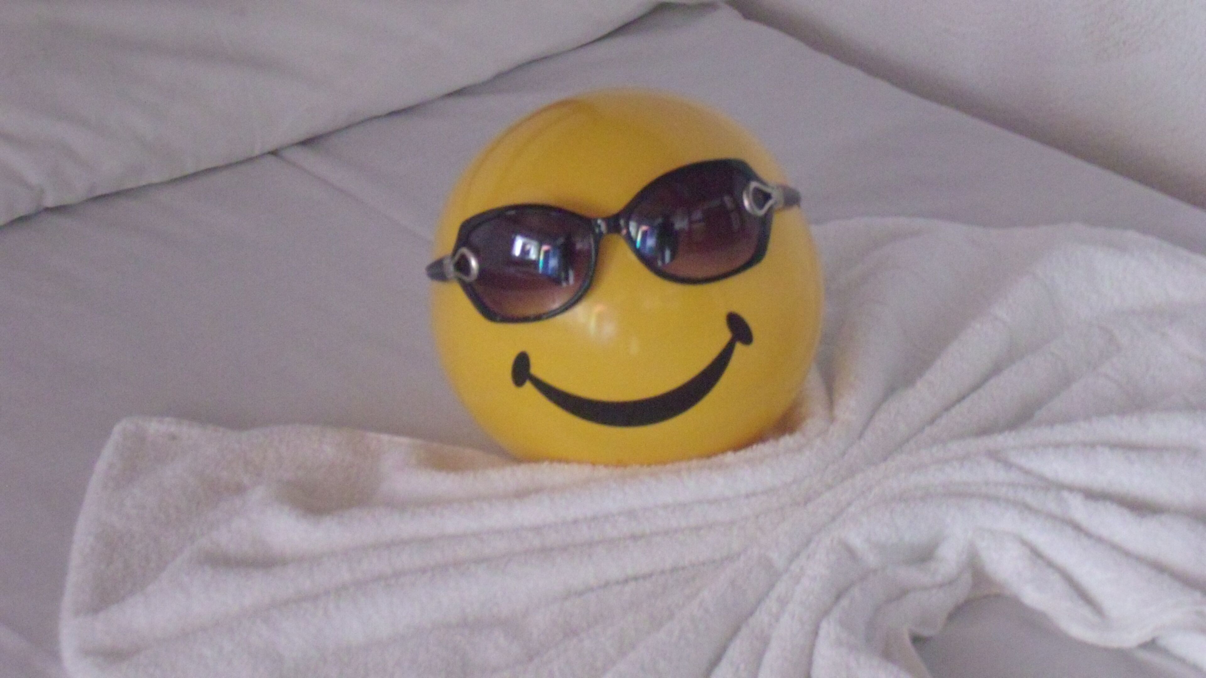 Ein gelbes Objekt mit Sonnenbrille und einem Smiley auf einem Bett mit weißer Bettwäsche.