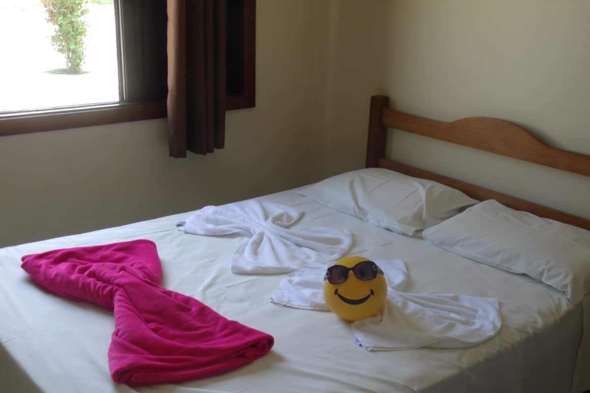 A bed with white linens, a pink towel, and a yellow plush toy with sunglasses.