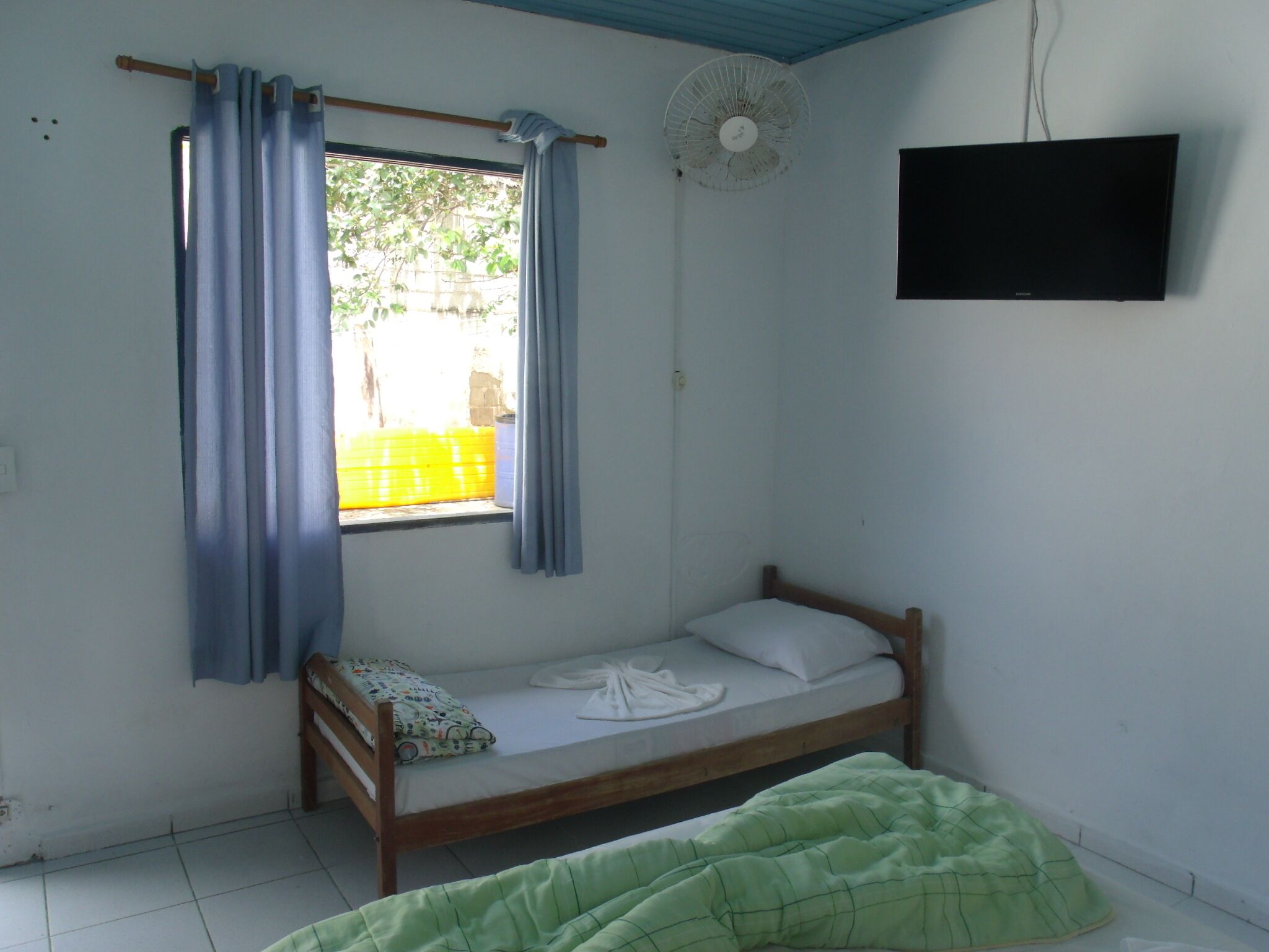 A small, single-bed room with a wooden frame, a green blanket, a white pillow, a window with blue curtains, a ceiling fan, and a TV mounted on the wall.