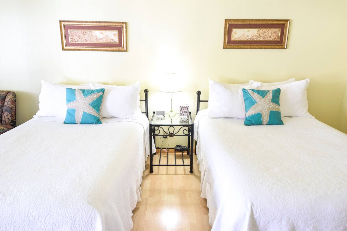 A hotel room with two beds, each with a white bedspread and a teal star-shaped pillow. There is a small table with a lamp between the beds, and framed artwork on the walls.