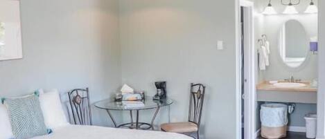 Quarto com cama grande, mesa pequena, cadeiras e banheiro com espelho e pia.