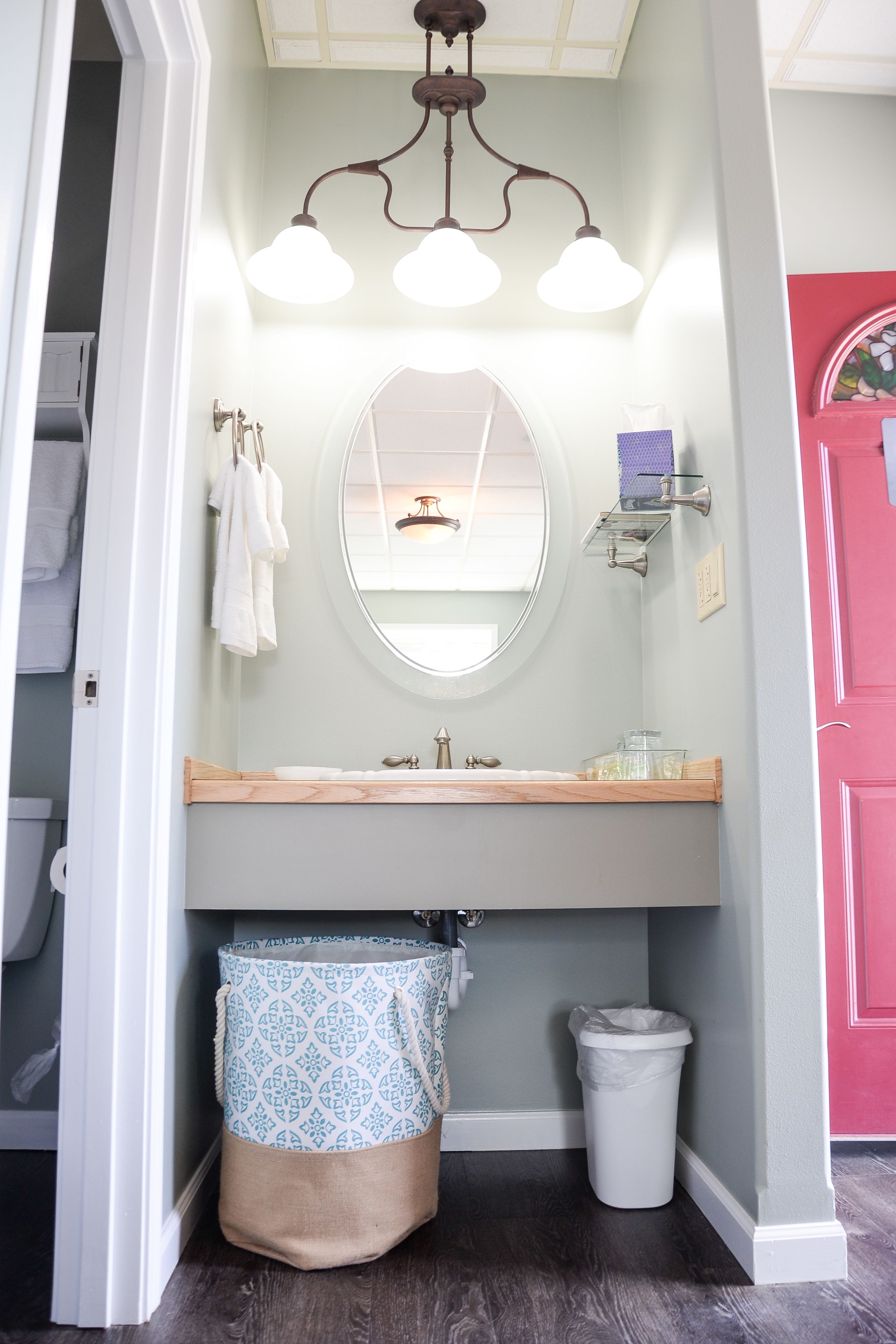 A bathroom with a round mirror, a light fixture with three bulbs, a sink, and a trash can.