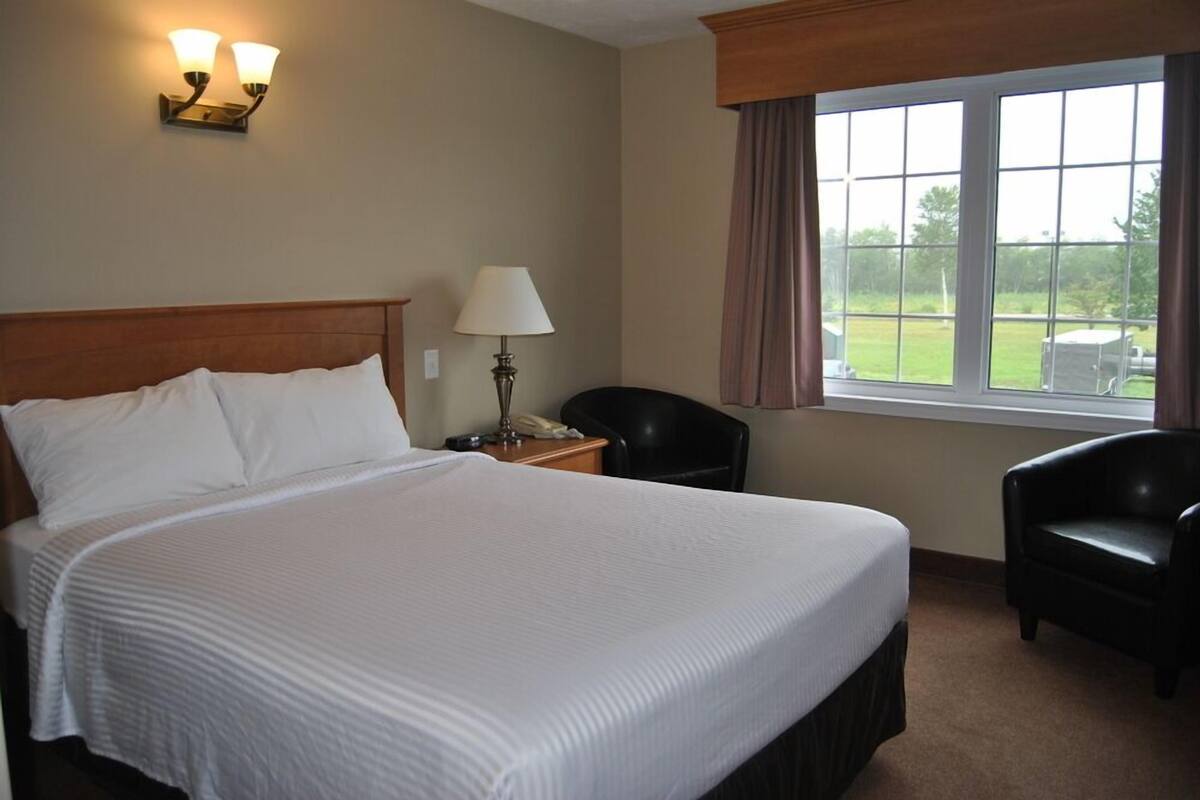Una habitación de hotel con una cama grande, una mesita de noche con lámpara, una silla y una ventana con cortinas.