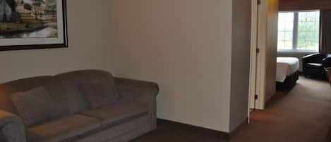 A room with a grey sofa, a framed landscape painting, and a bed with a white comforter.