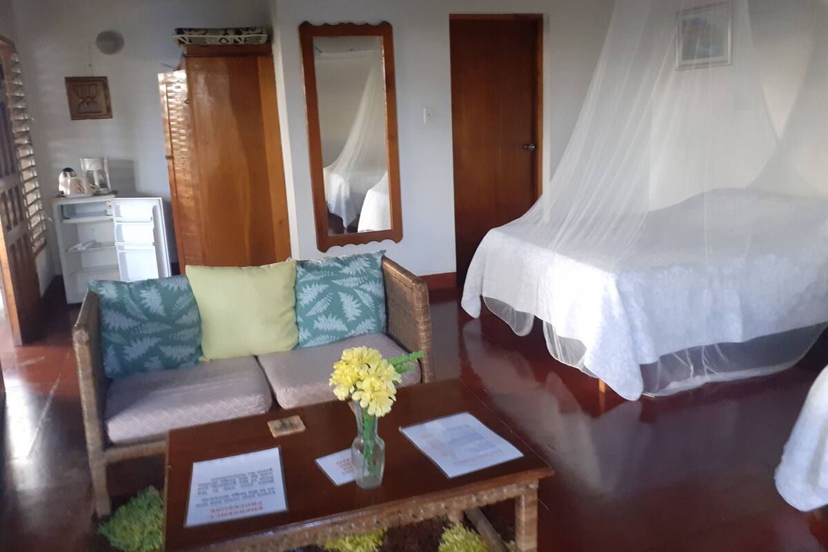 A room with a bed, a sofa, a small table with papers, a mirror, and a refrigerator.