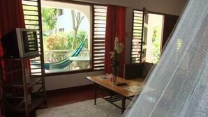 A room with a wooden table, a TV on a stand, a window with red curtains, and a view of greenery outside. - LTU Garden (Negril)