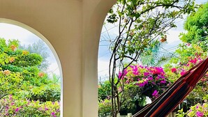 A hammock under a covered patio with a view of a garden with pink flowers and green foliage. - LTU Garden (Negril)