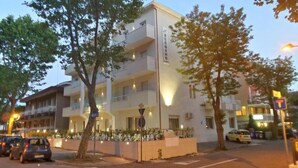 Un hôtel à plusieurs étages, dont la façade porte une enseigne indiquant « BERGAMO ». Des arbres se trouvent devant l’immeuble, et une rue bordée de voitures garées et d’un panneau de signalisation est visible.