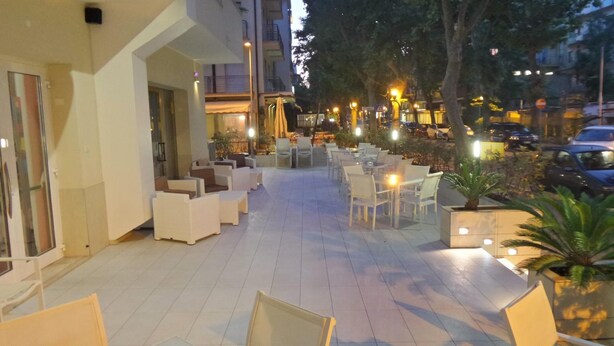 Un espace extérieur aménagé avec des tables et des chaises, entouré de plantes en pot et bordé par la façade d’un bâtiment.
