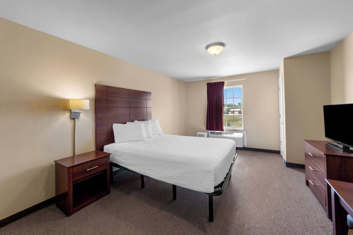 Une chambre d’hôtel avec un lit, une table de chevet, une lampe, une télévision et une fenêtre avec des rideaux.