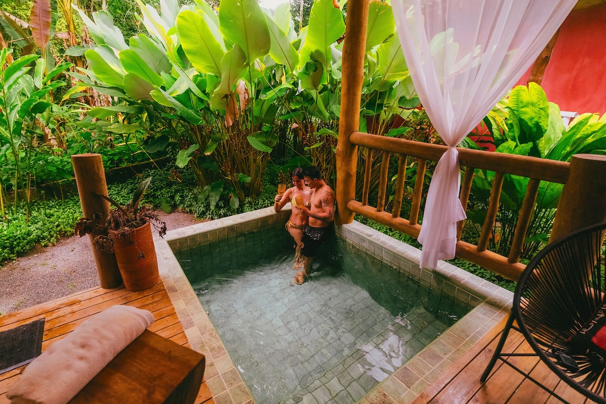 A small, private hot tub surrounded by lush greenery, with a wooden deck and a wicker chair nearby.