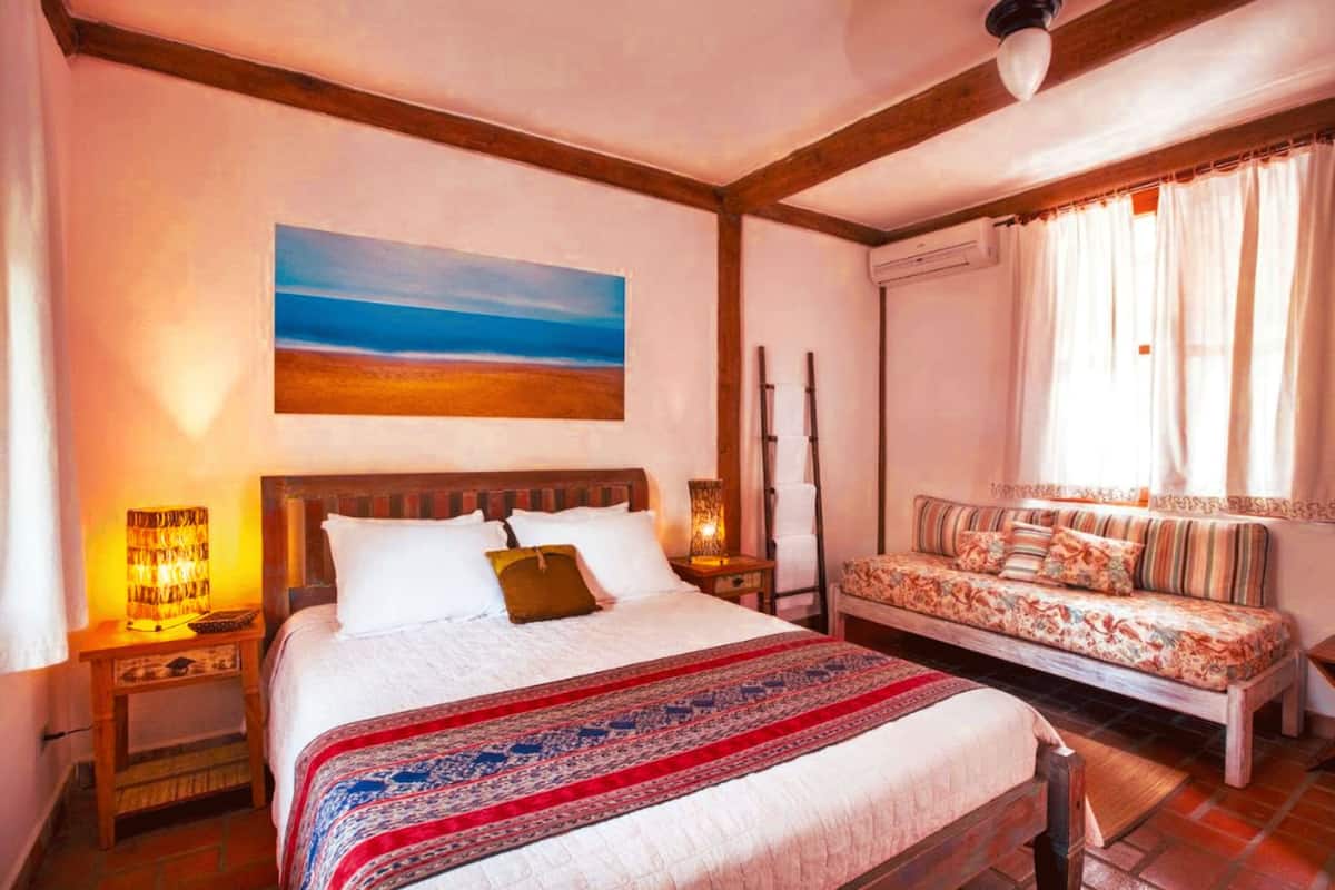 A bedroom with a large bed, a sofa, a painting of a beach, and a window with curtains.
