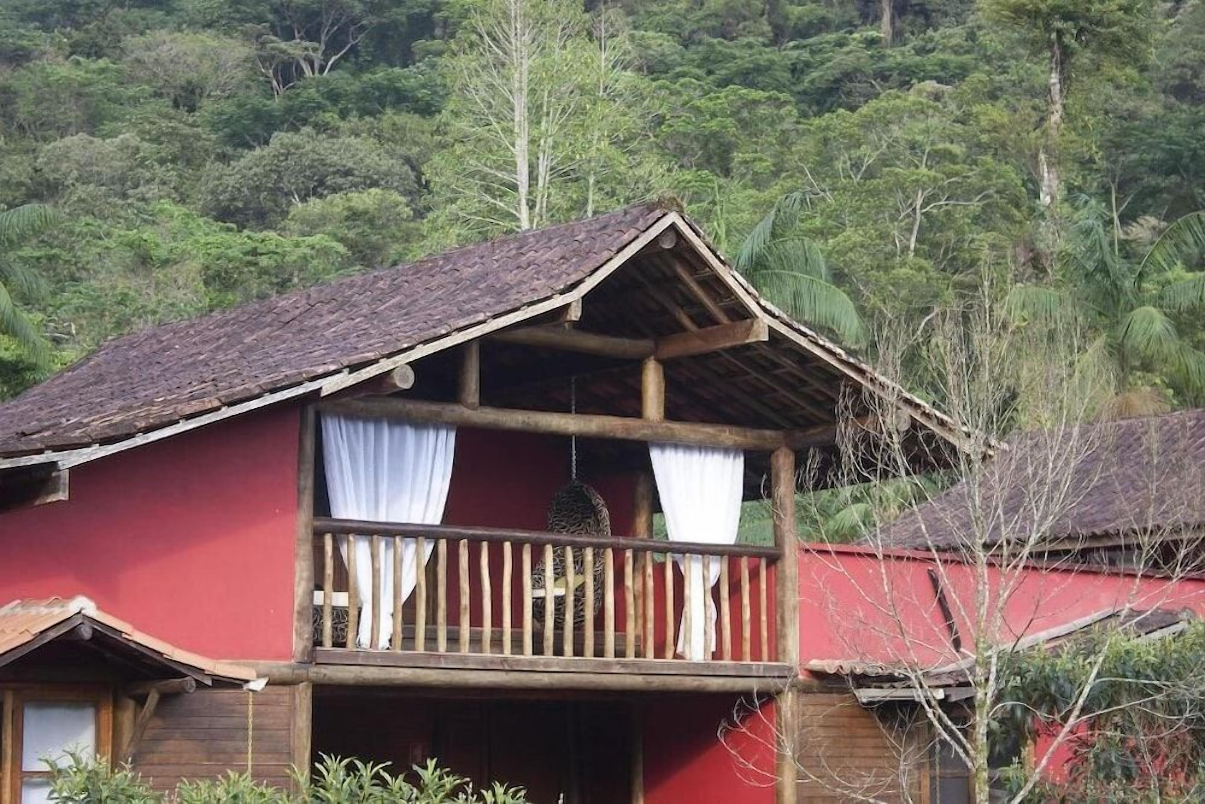 Uma casa de madeira de dois andares com varanda, rodeada por vegetação exuberante.
