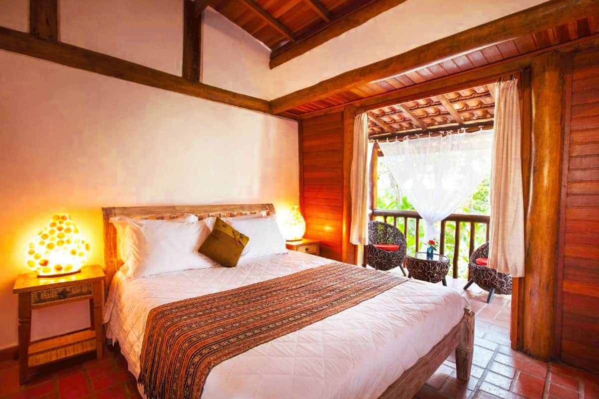 A bedroom with a large bed, wooden beams, a balcony with chairs, and a view of greenery.