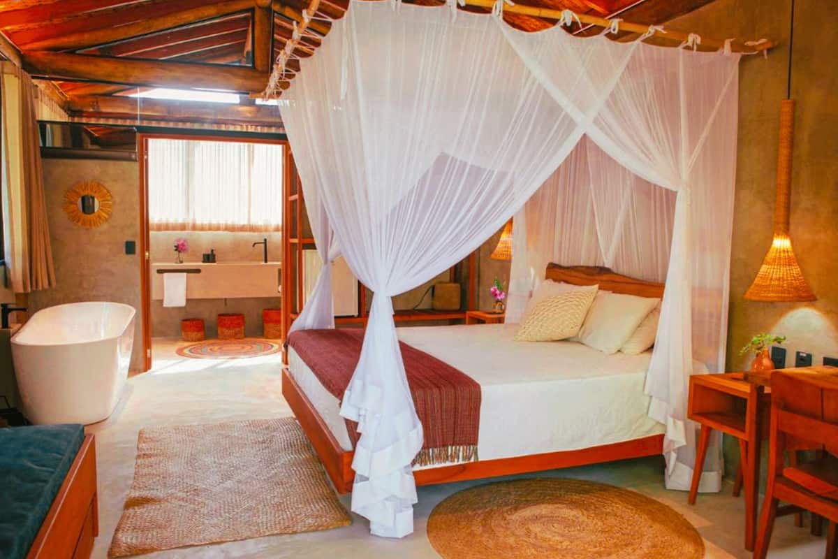A bedroom with a canopy bed, a sitting area with a chair and table, a bathroom with a bathtub, and a small desk with a lamp.