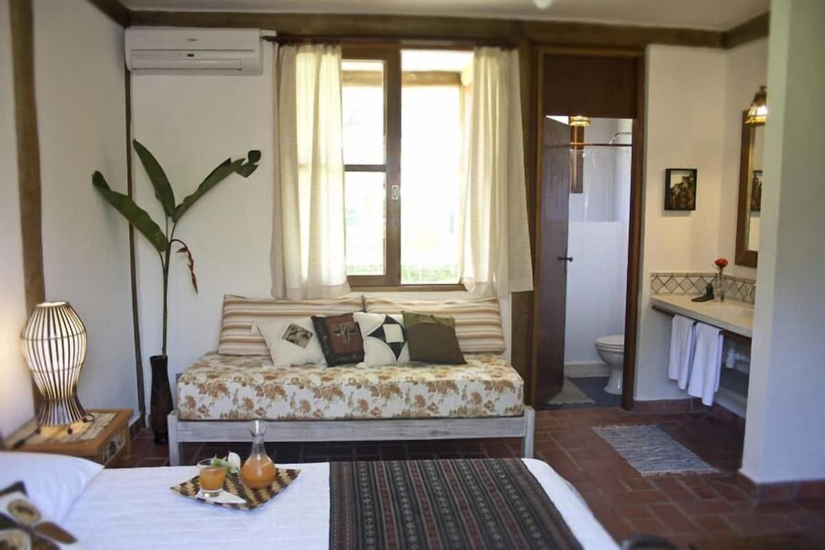 A bedroom with a bed, a bedside table with a lamp, a window with curtains, a bathroom with a sink and mirror, and a plant.