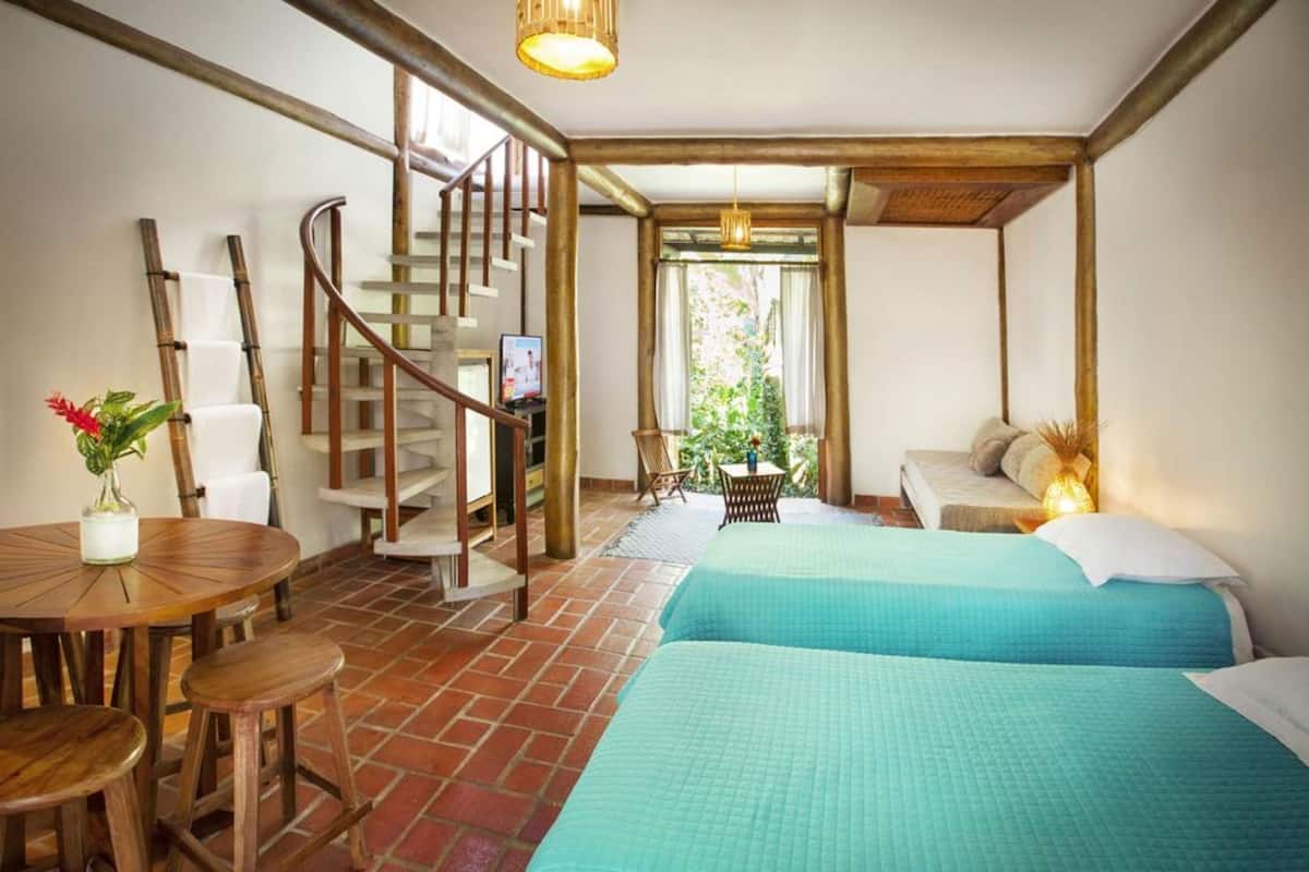 A room with a spiral staircase, two beds with blue bedding, a round wooden table with a vase of flowers, and a small table with a plant.
