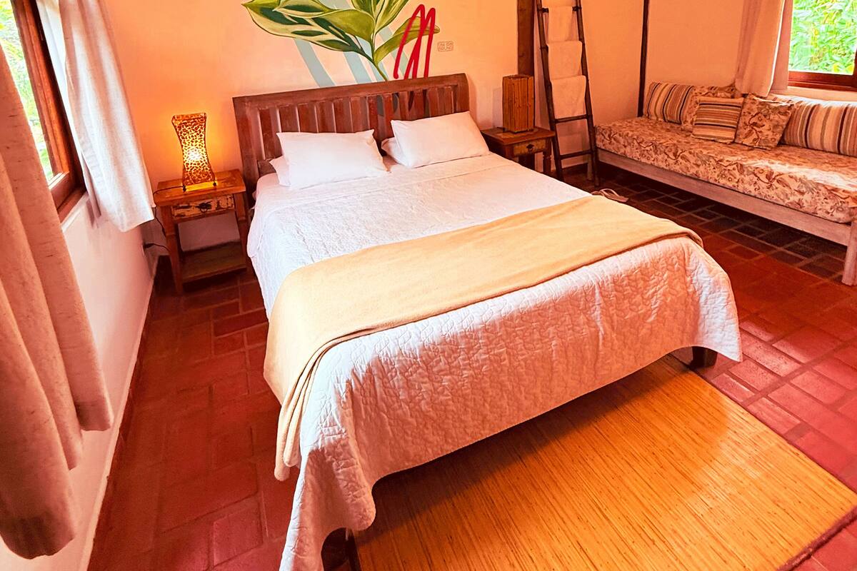 A neatly made bed with a wooden headboard, a bedside table with a lamp, a bench with cushions, and a large plant.
