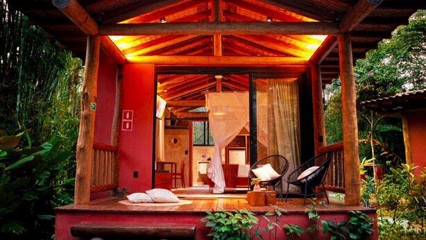 Une structure en bois à l’extérieur rouge, dotée d’un porche couvert et offrant une vue sur une pièce où se trouvent un lit et une chaise.