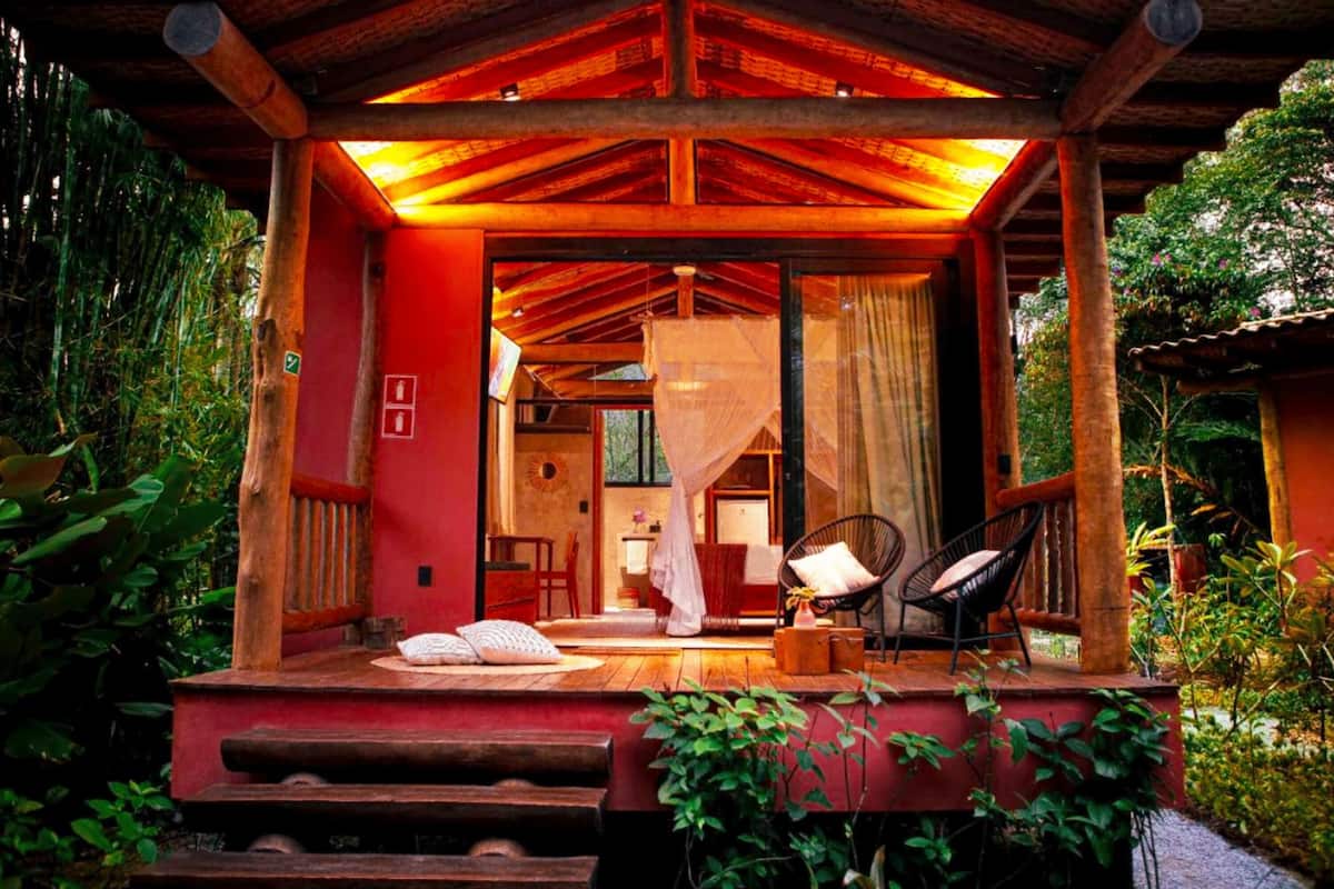 A wooden structure with a red exterior, a covered porch, and a view into a room with a bed and a chair.