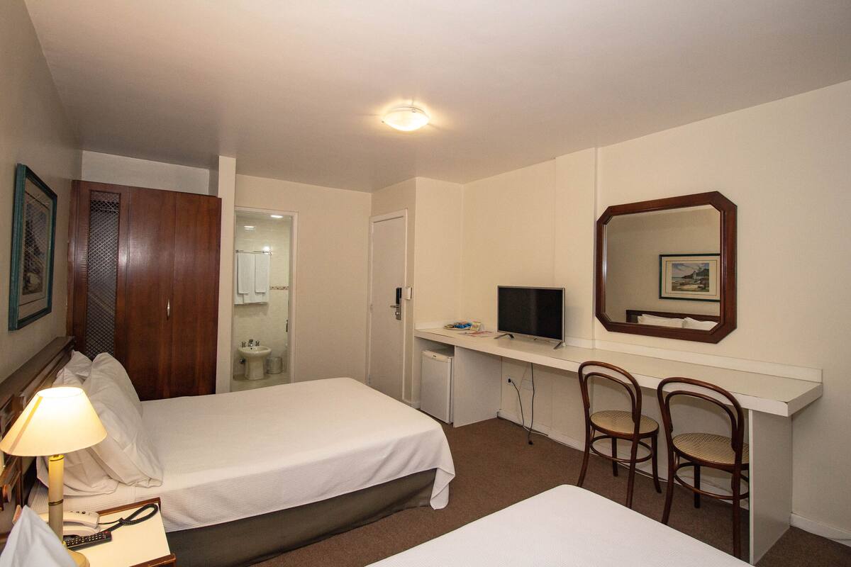 Une chambre d’hôtel avec deux lits, un bureau équipé d’un ordinateur et d’une chaise, une télévision et une salle de bain visible à travers une porte ouverte.