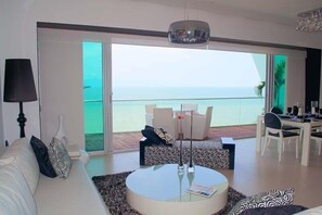 A modern living room with a large balcony overlooking the sea, a round coffee table, and a dining area with white furniture.