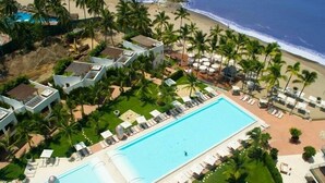 Et resort med en stor swimmingpool, flere loungeområder og udsigt til stranden.
