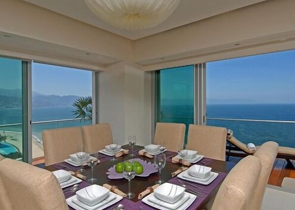 A dining area with a table set for six, large windows with a view of the sea, and a ceiling fan.