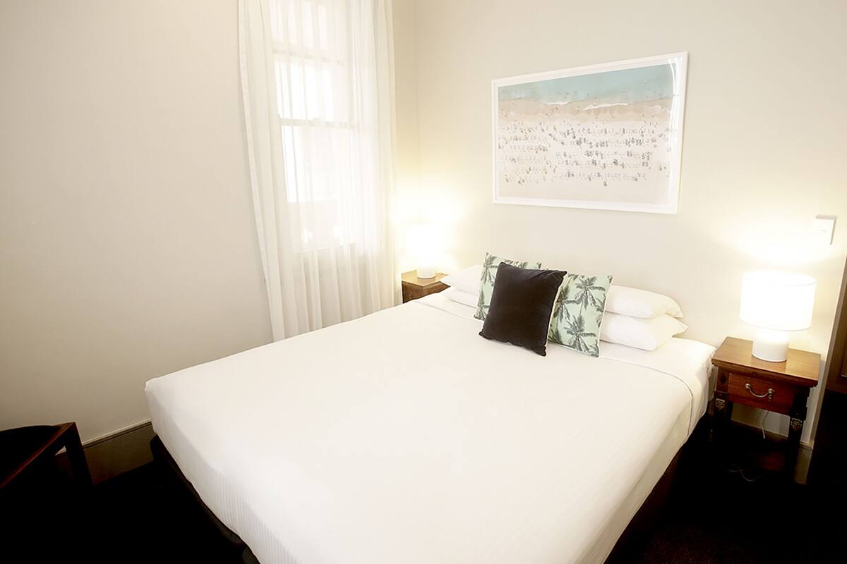 A hotel room with a large bed, two bedside tables with lamps, a window with sheer curtains, and a beach-themed wall art.