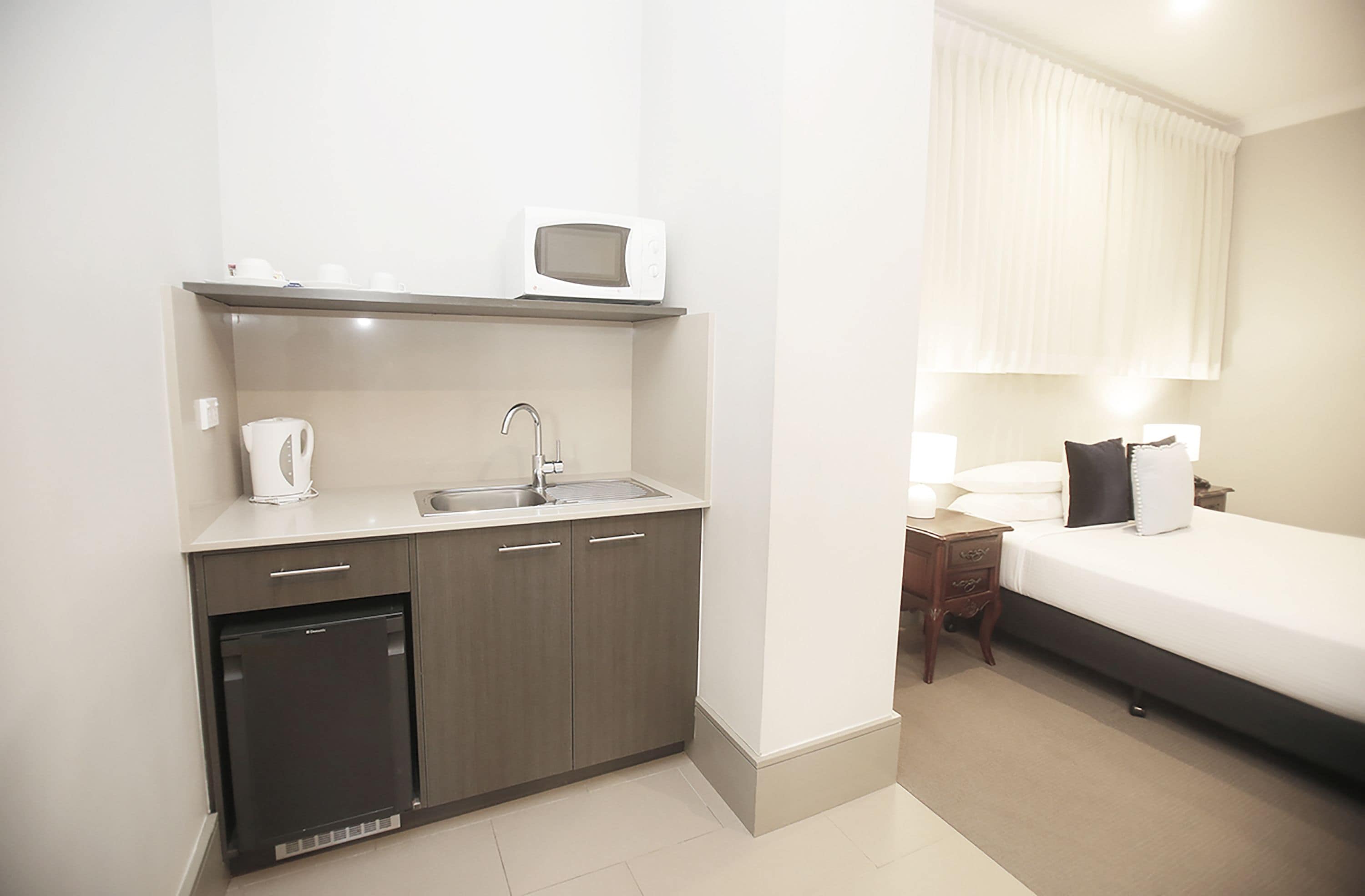 A compact hotel room with a kitchenette, a bed, and a bedside table.
