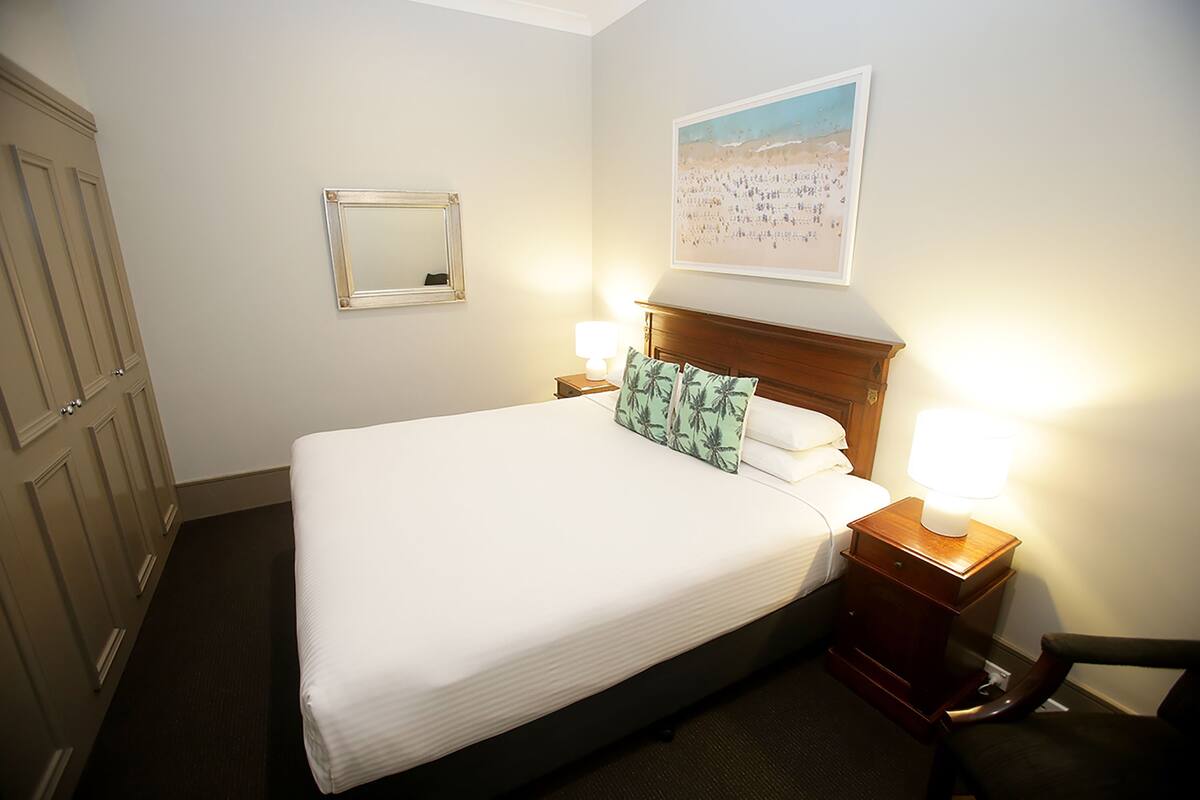 A neatly made bed with a wooden headboard, two bedside lamps, a framed mirror, and a beach-themed wall art.
