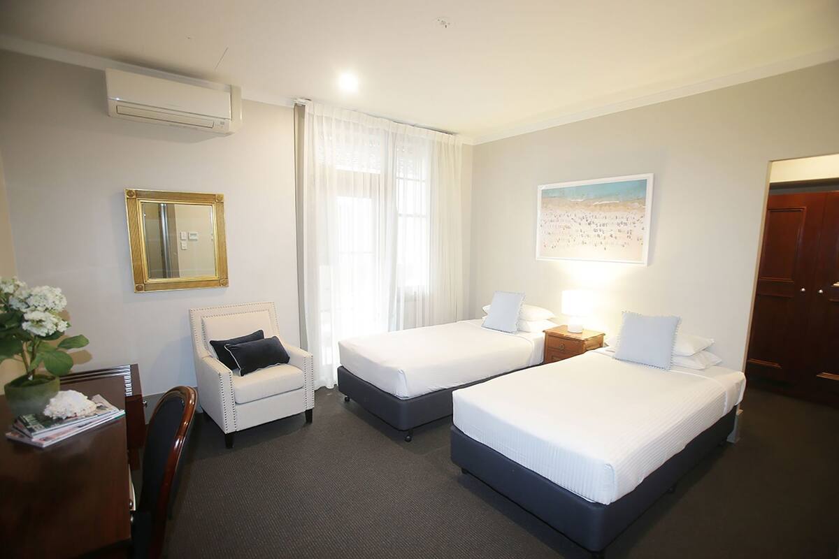A hotel room with two beds, a desk with a chair, a potted plant, a mirror, and a painting on the wall.