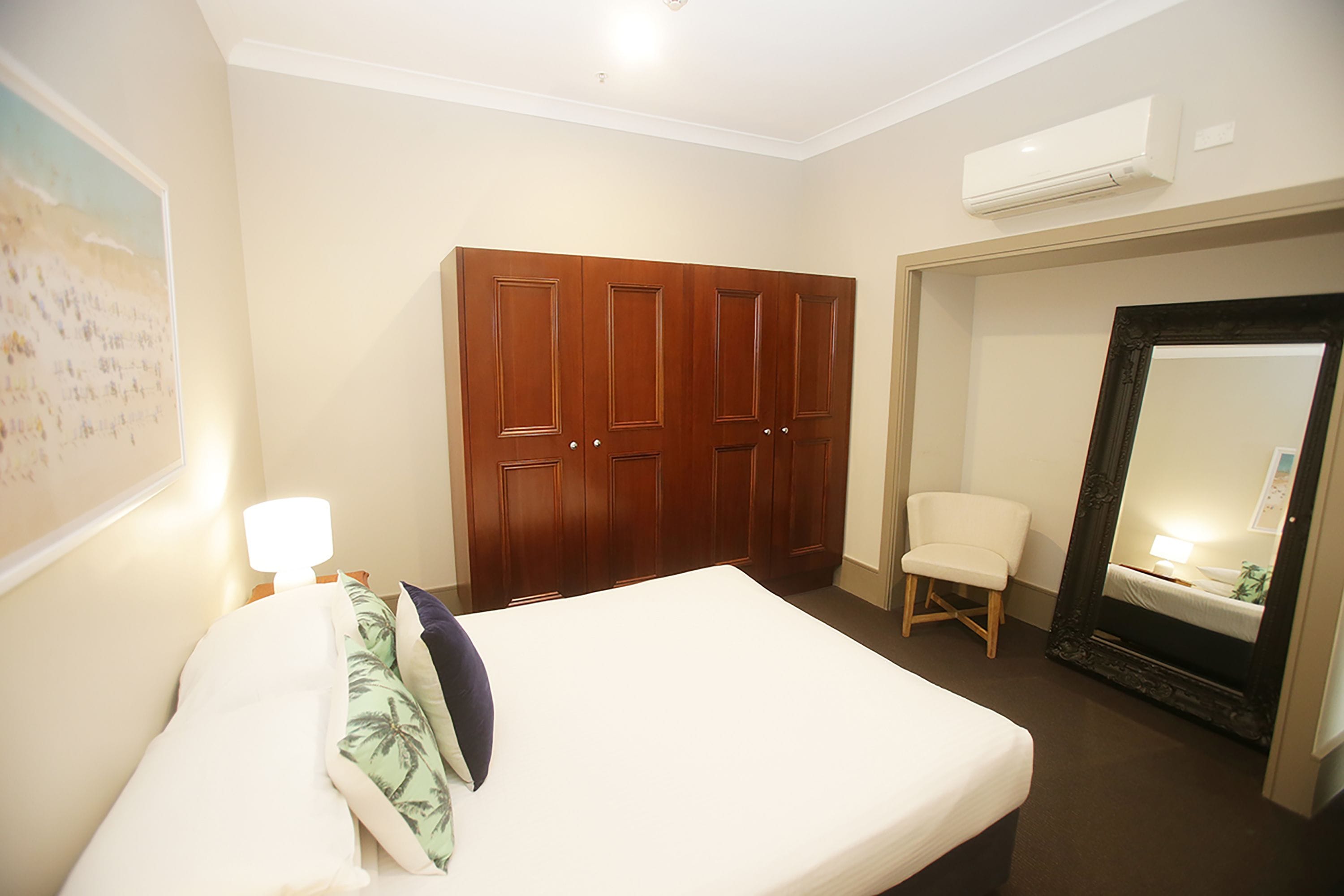 A hotel room with a bed, a wooden wardrobe, a chair, a large mirror, and an air conditioning unit.