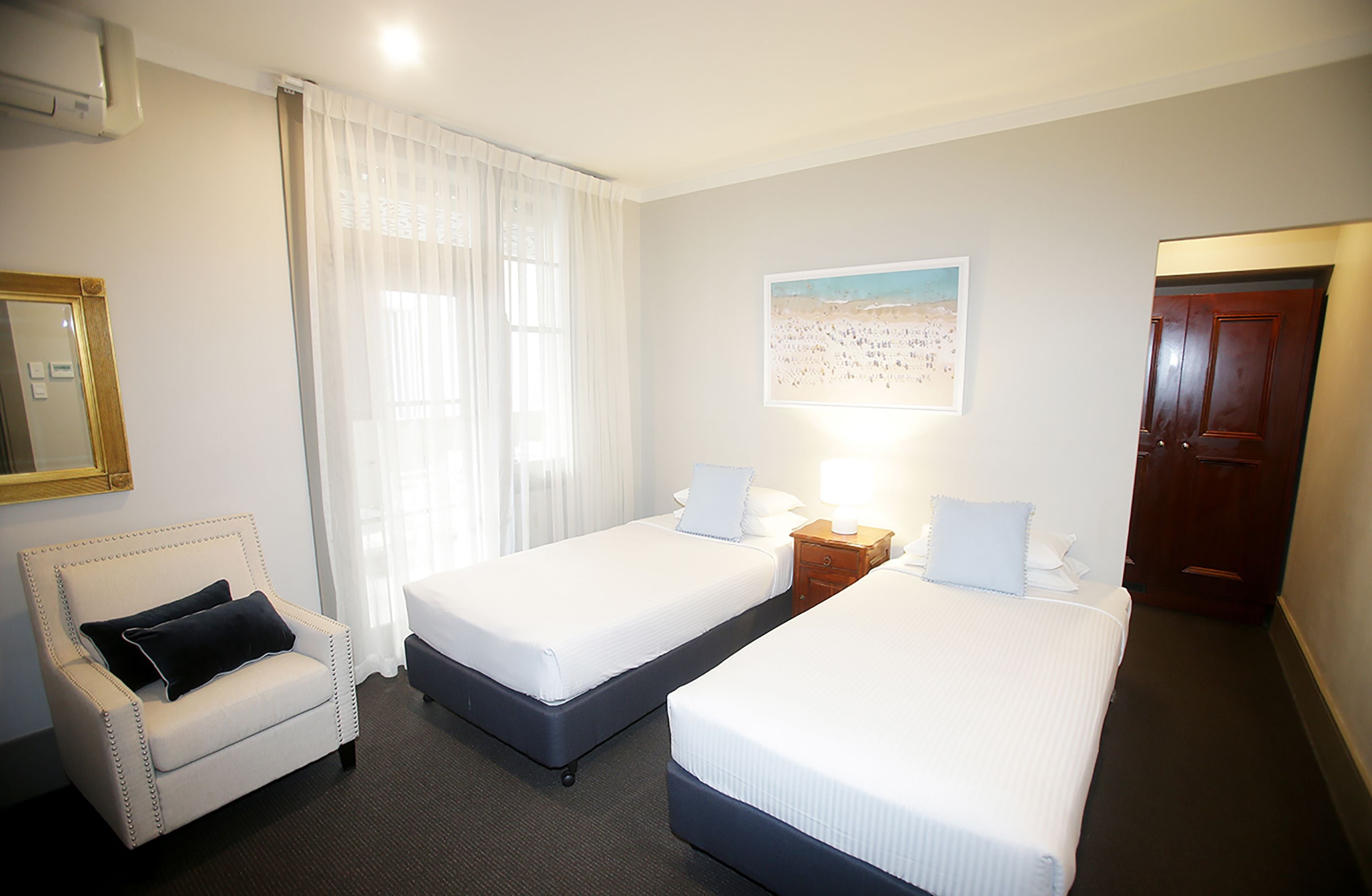A hotel room with two single beds, a chair, a nightstand, a lamp, a painting on the wall, and a door.