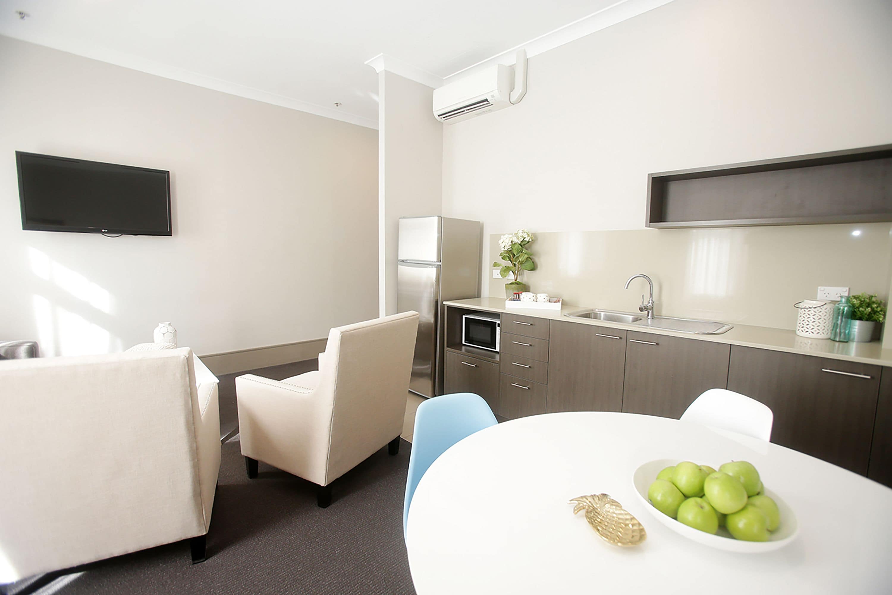 A modern hotel room with a flat-screen TV, a dining area with a white table and chairs, a kitchen with a refrigerator and microwave, and a sink.