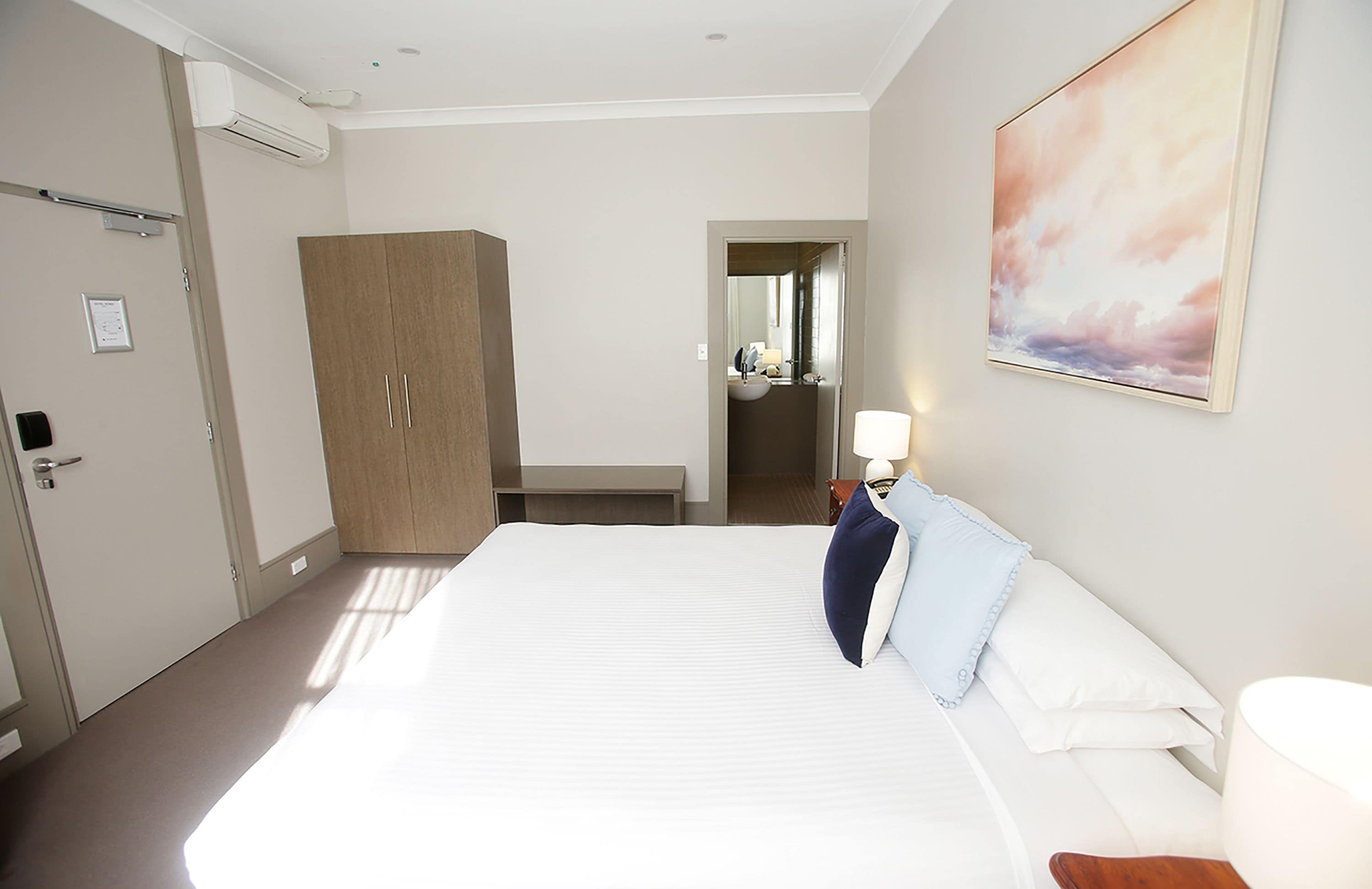 A hotel room with a large bed, a wooden wardrobe, a bathroom visible through an open door, and a painting on the wall.