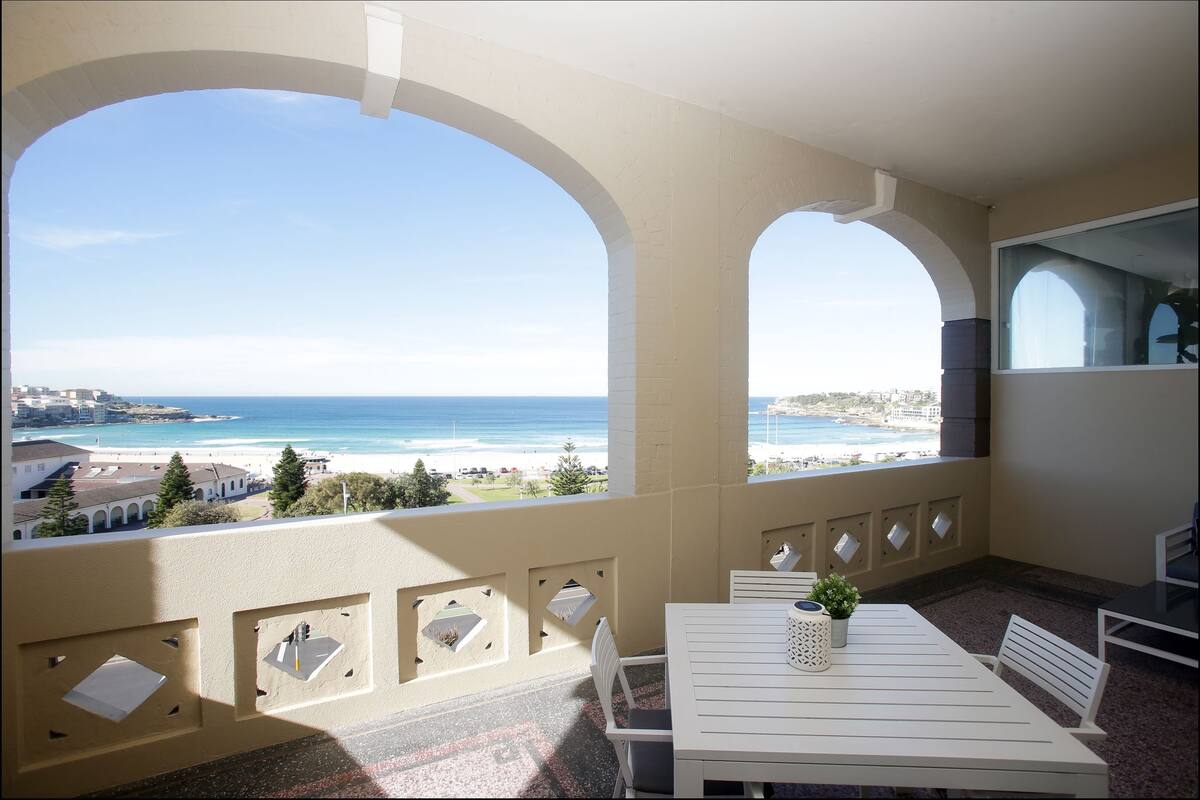 A balcony with a table and chairs, overlooking a beach and ocean.