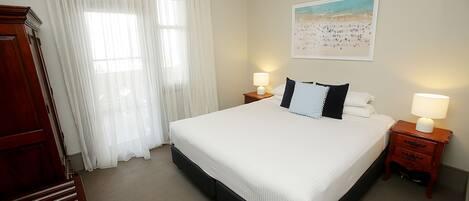 A neatly made bed with white linens, two bedside tables with lamps, a wooden nightstand, a large window with sheer curtains, and a beach-themed painting on the wall.