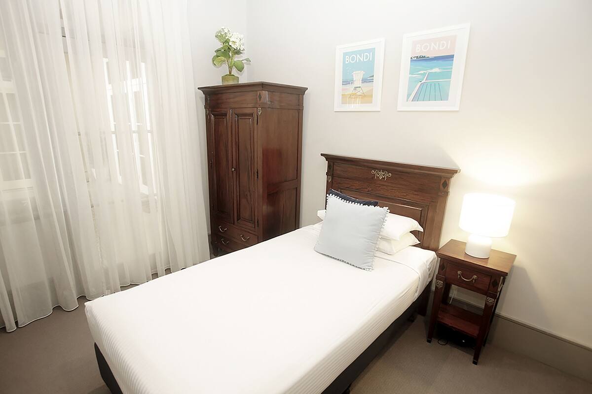 A neatly made bed with a white pillow and a wooden headboard, a small bedside table with a lamp, a wooden wardrobe, and two framed pictures on the wall.