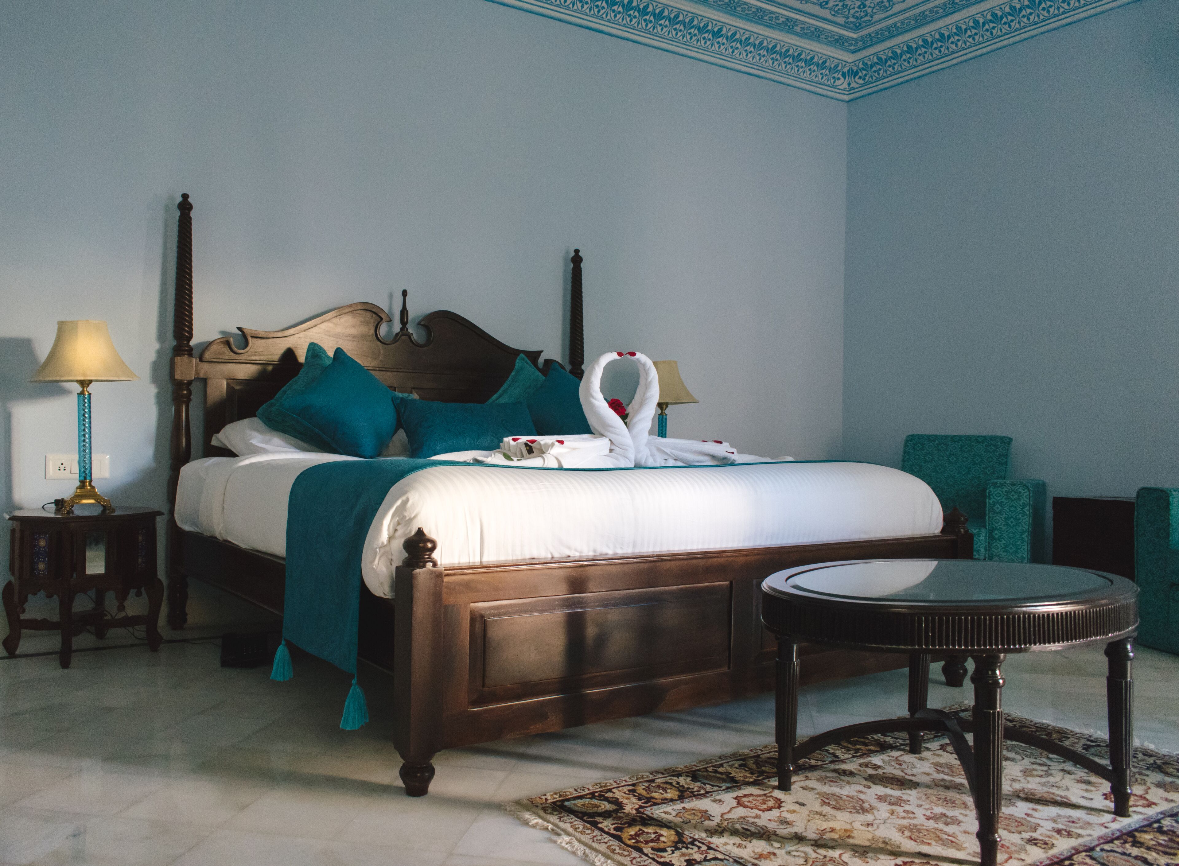 A neatly made bed with a wooden headboard, teal pillows, and a swan-shaped towel arrangement. There is a small round table with a patterned rug beside the bed.