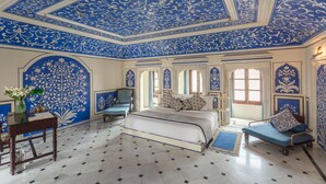Une pièce comprenant un grand lit, un bureau et une chaise. Les murs et le plafond sont ornés de motifs décoratifs.