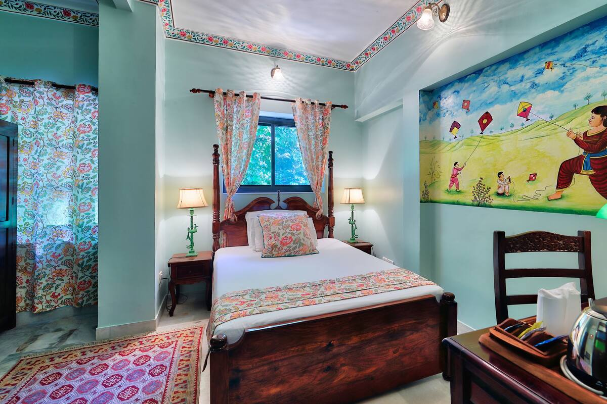 Une chambre avec un grand lit, deux tables de chevet, une lampe verte, un tableau représentant un festival de cerfs-volants et une vue sur l’extérieur.