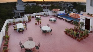 Terraza en la azotea con varias mesas y sillas, rodeada de plantas en macetas, y un área de piscina con sombrillas.