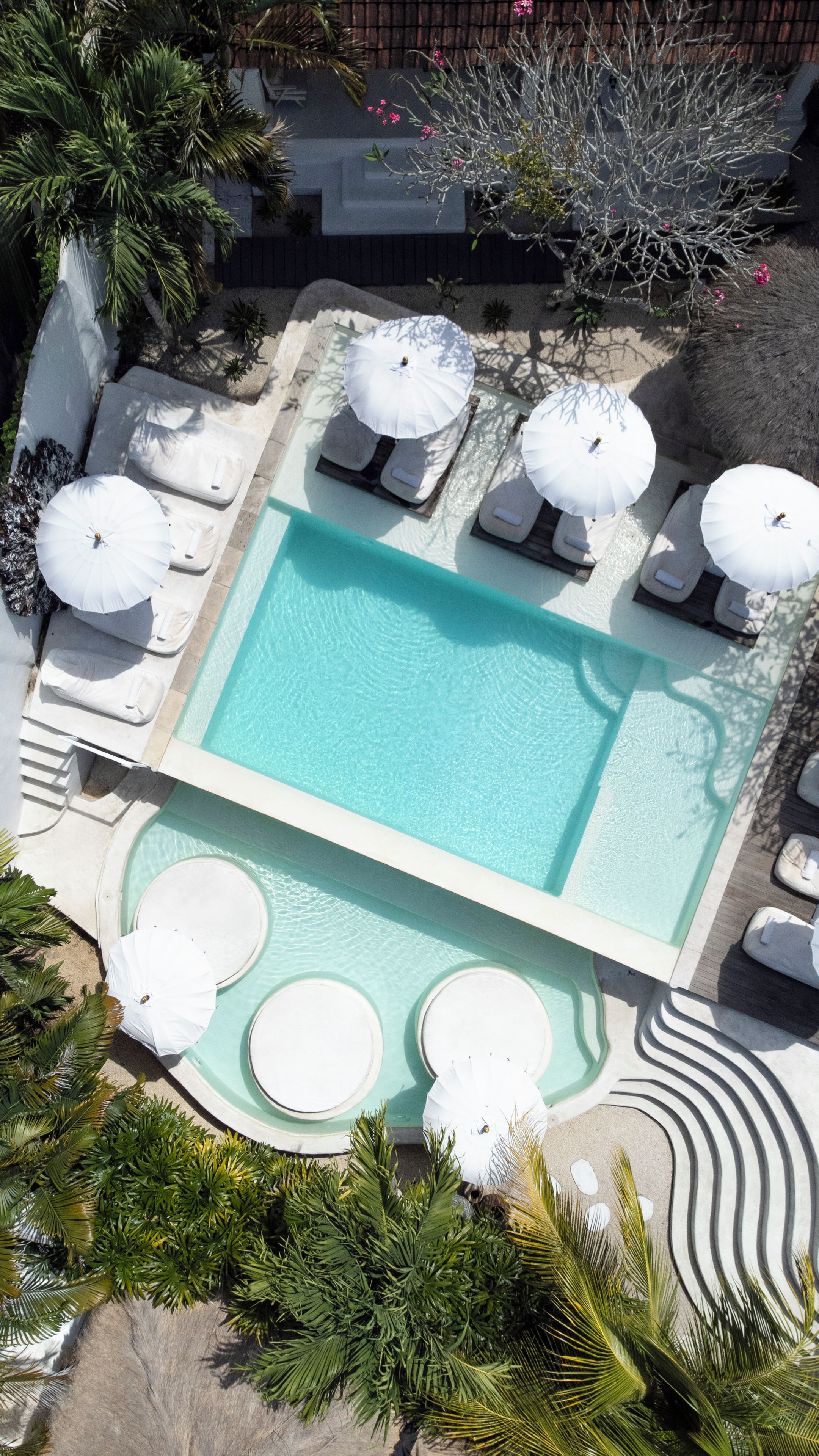 An aerial view of a pool area with lounge chairs and umbrellas, surrounded by palm trees and a wooden deck.