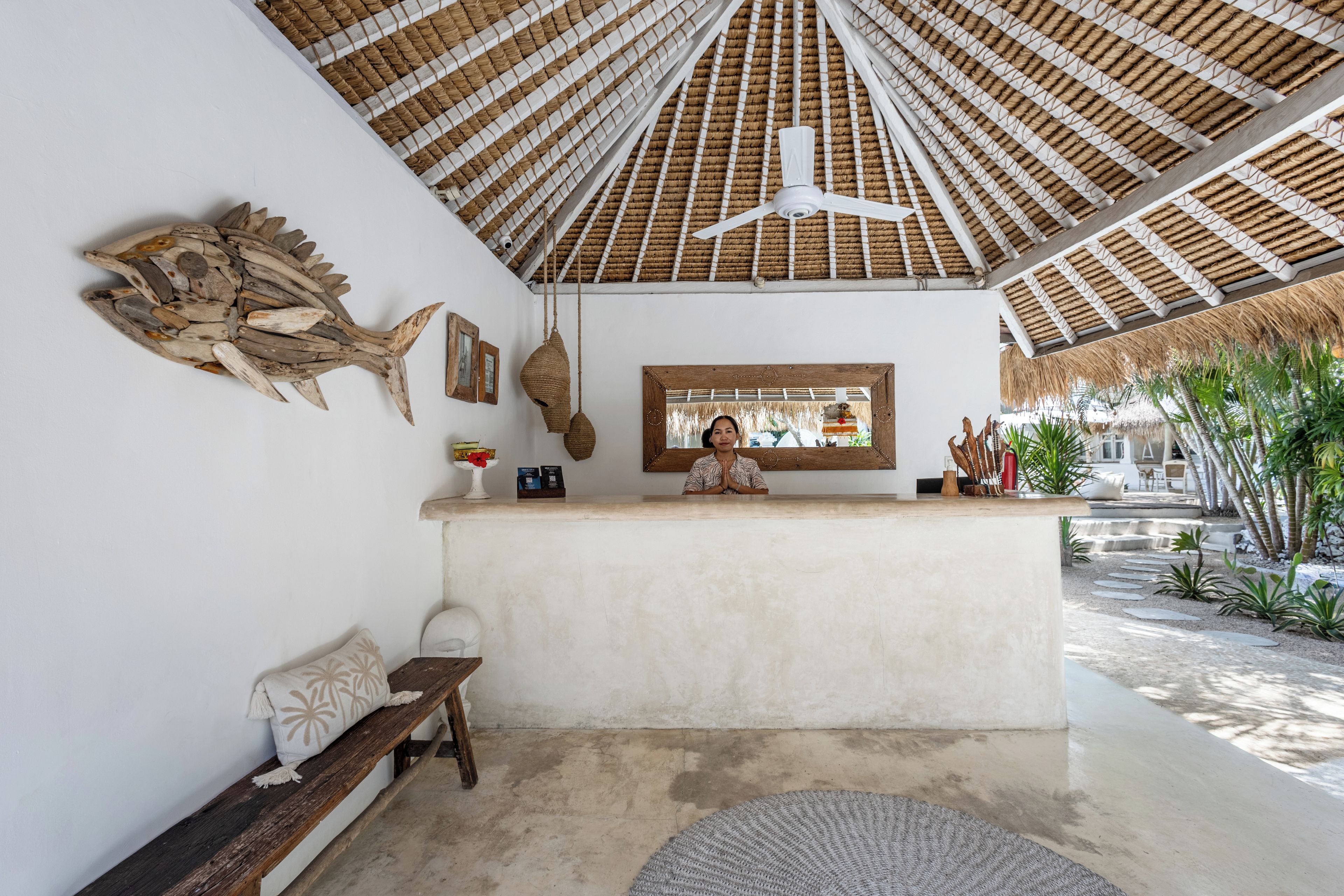 A reception area with a thatched ceiling, a wooden bench, a framed picture, and a small table with a red flower vase.