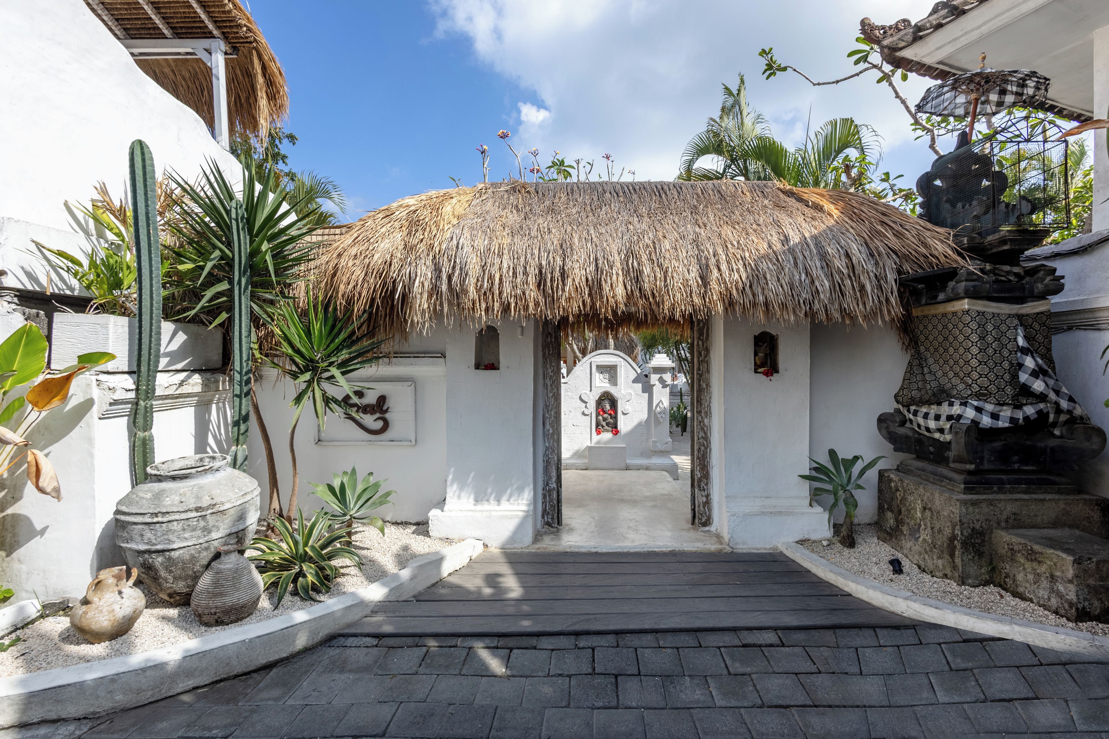 A pathway leading to a building with a thatched roof, surrounded by tropical plants and a decorative fountain.