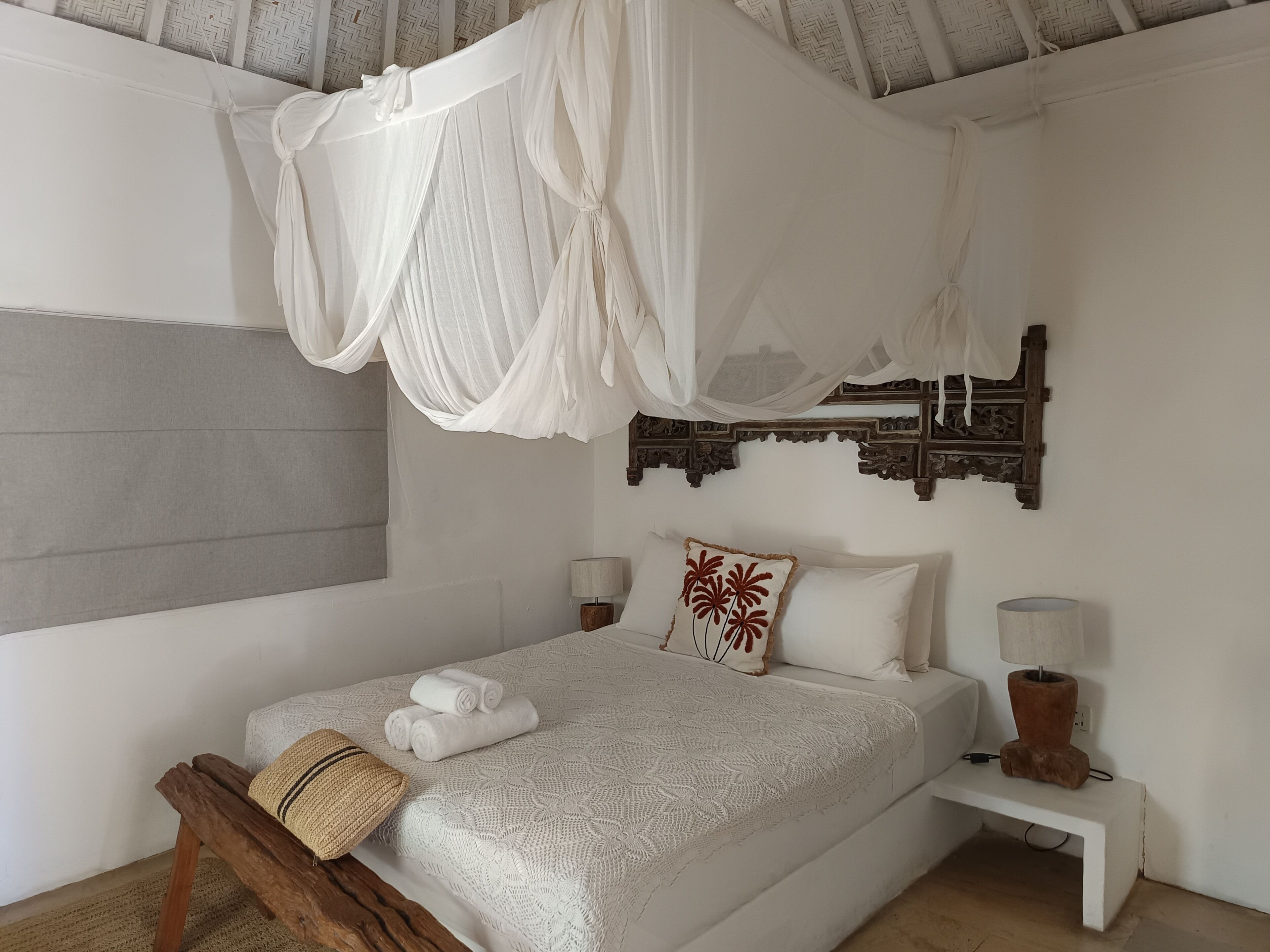 Een slaapkamer met een hemelbed, een geweven stoel, een nachtkastje met een lamp en een decoratief wandobject.