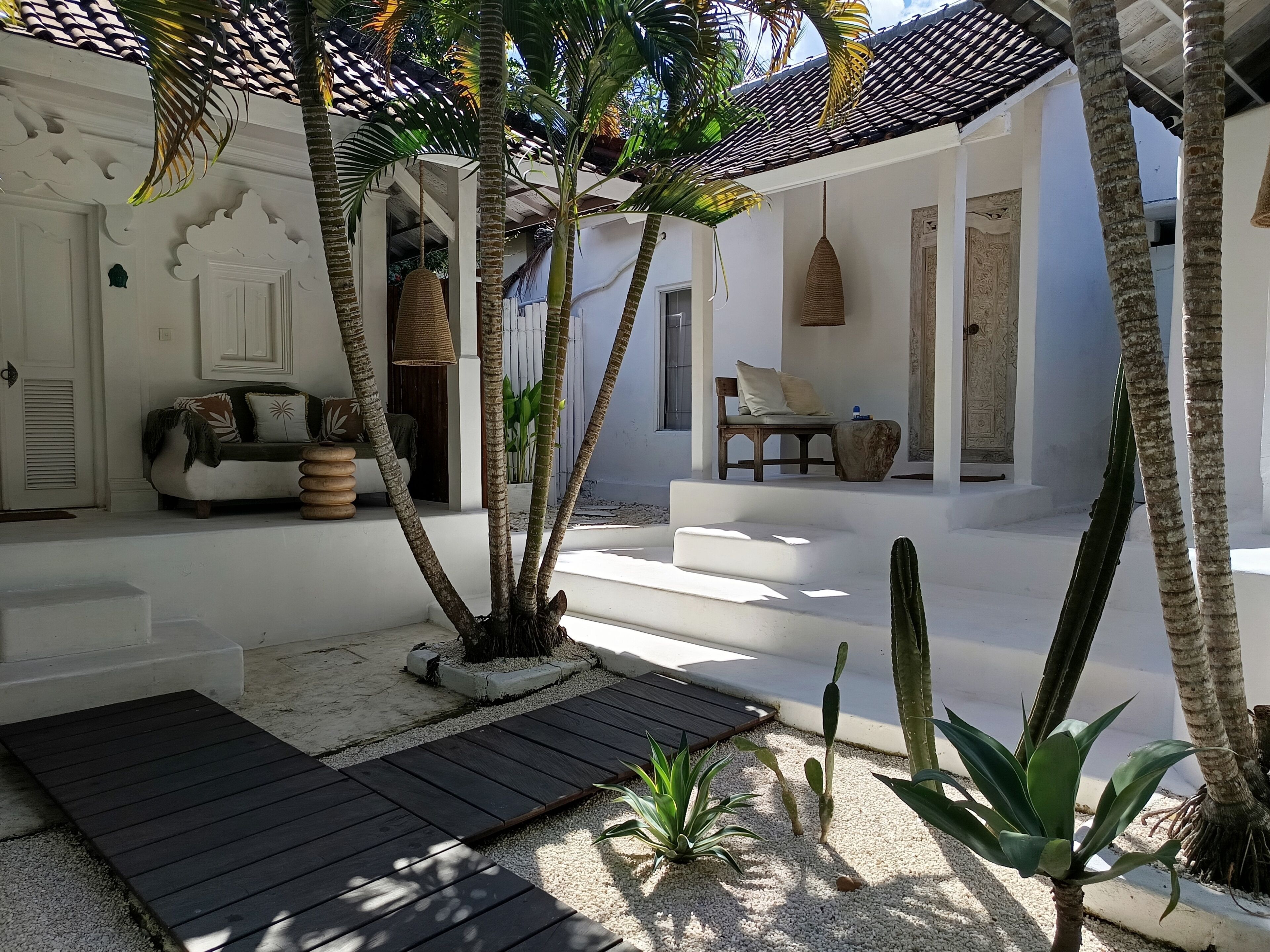 A white building with a covered patio, a wooden bench, a hanging light fixture, and a cactus plant.