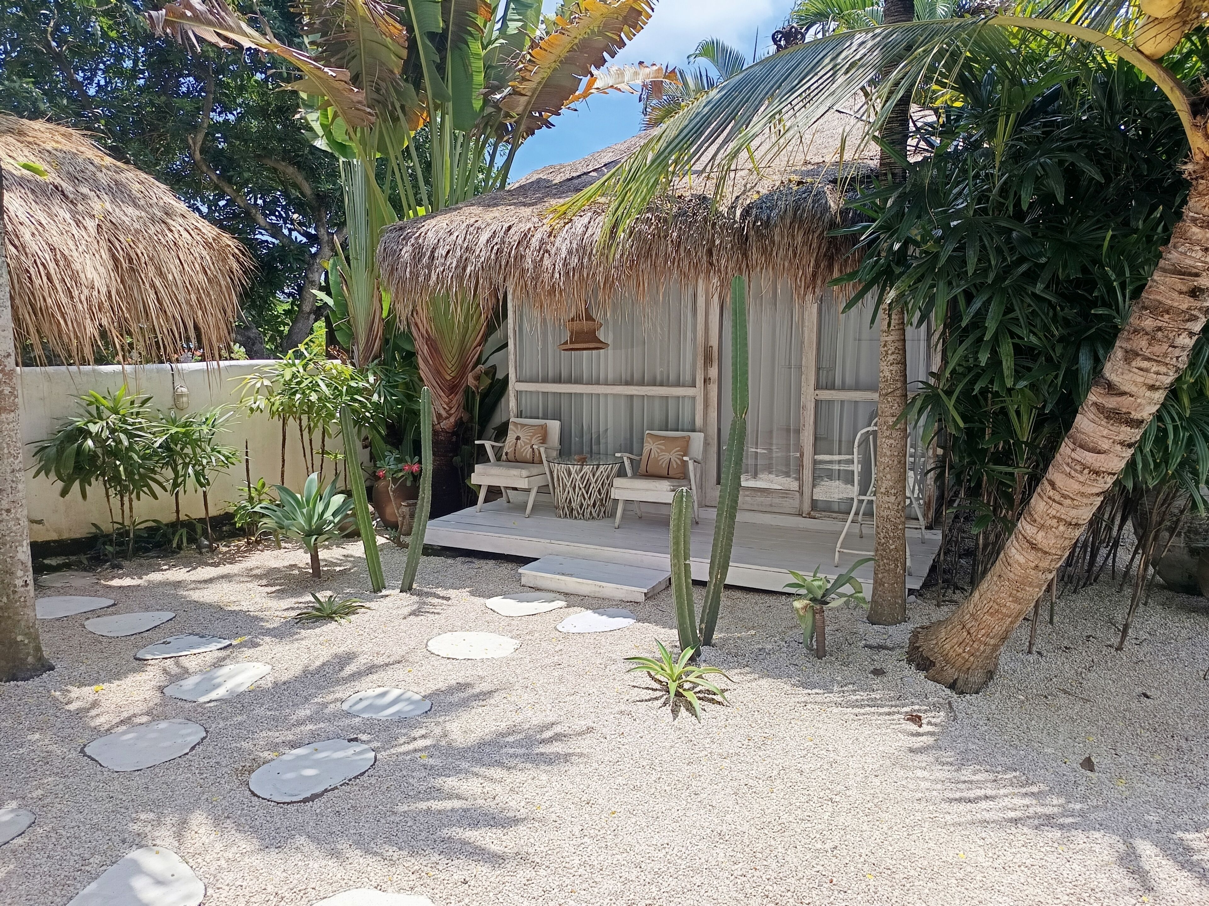 A tropical outdoor seating area with thatched-roof structures, palm trees, and a gravel pathway.