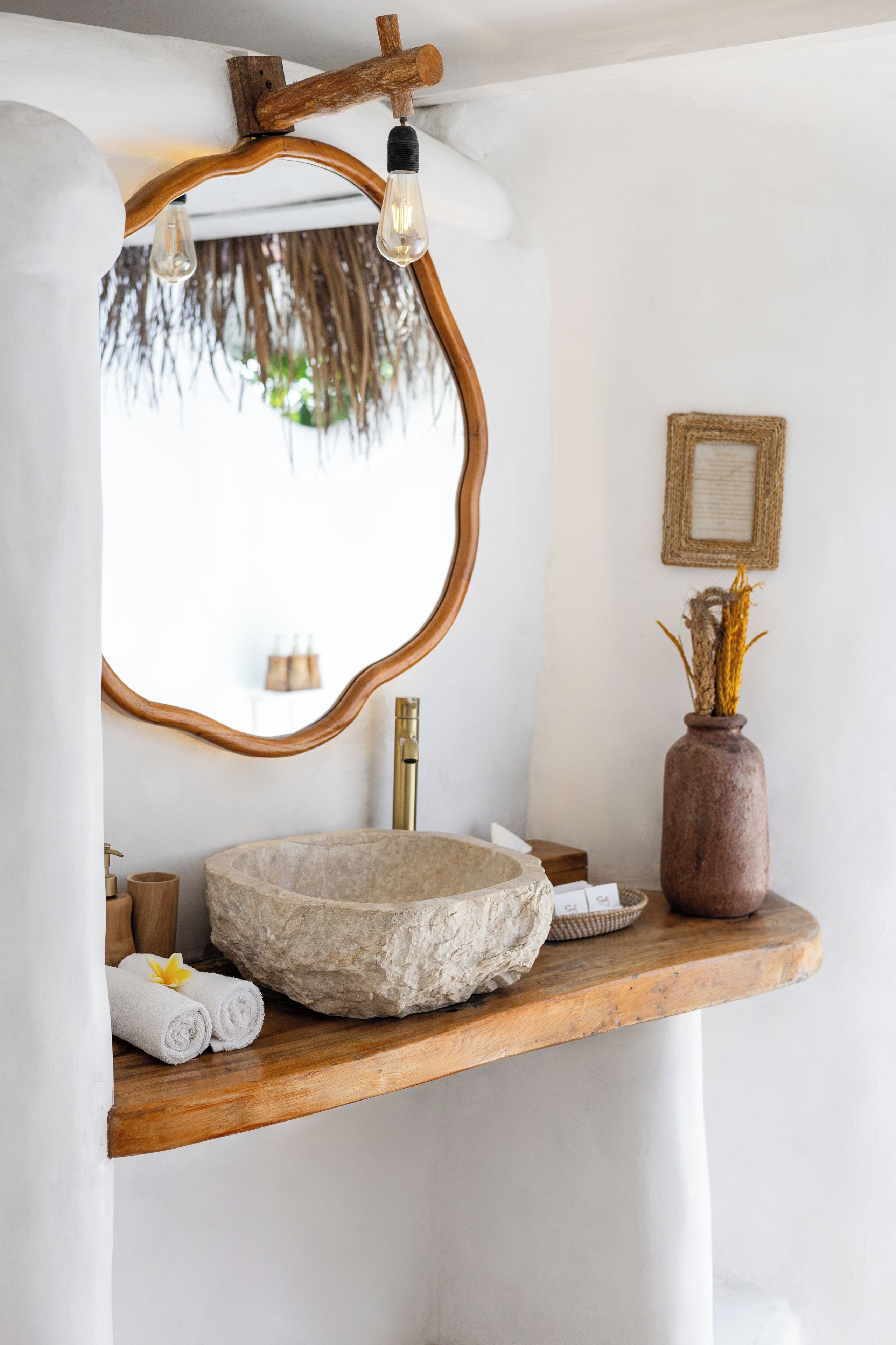 A bathroom with a stone sink, wooden vanity, a mirror with a wooden frame, a light fixture, and a vase with dried plants.